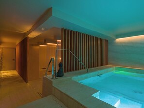 A modern spa area with a hot tub, wooden slats, and a person sitting by the tub. - San Vito Resort & Spa (San Vito Lo Capo)