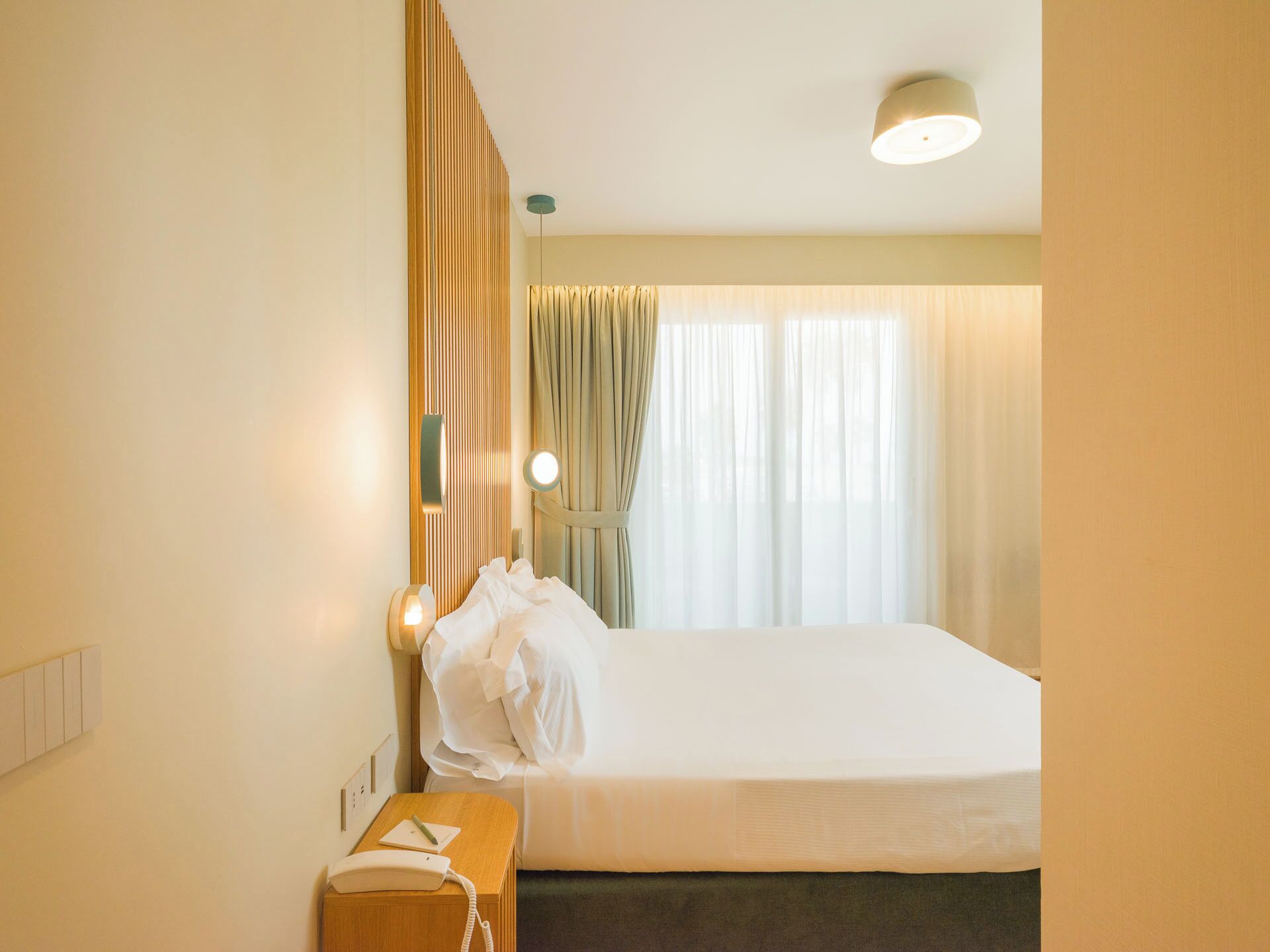 Ein Hotelzimmer mit einem Bett, einem Nachttisch mit Telefon, einer Lampe und einem Fenster mit Vorhängen.