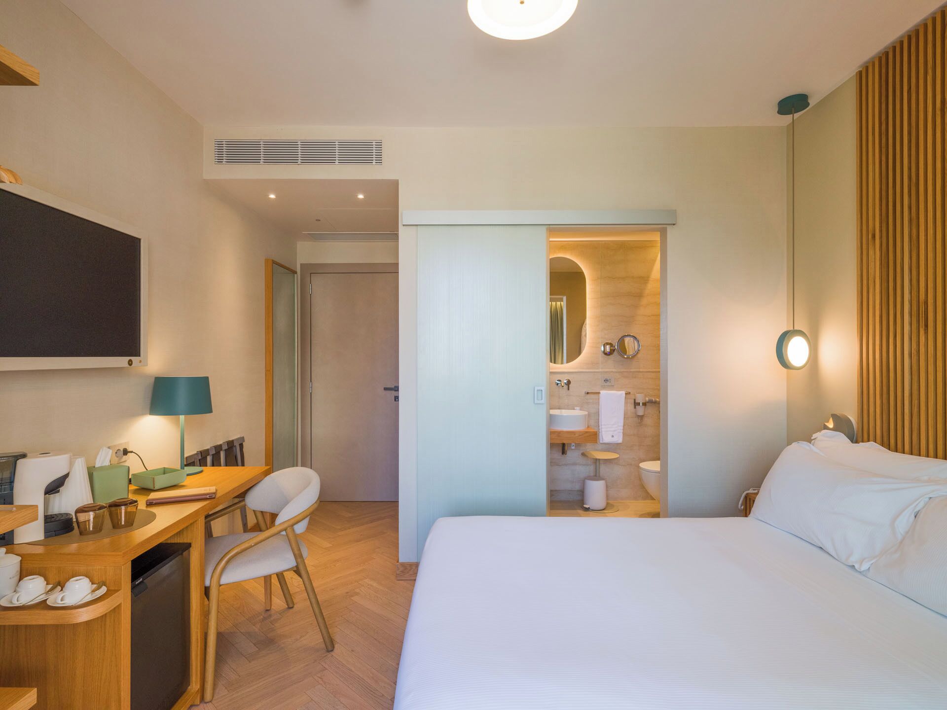 A hotel room with a bed, a desk, a chair, a mirror, a toilet, and a shower area.