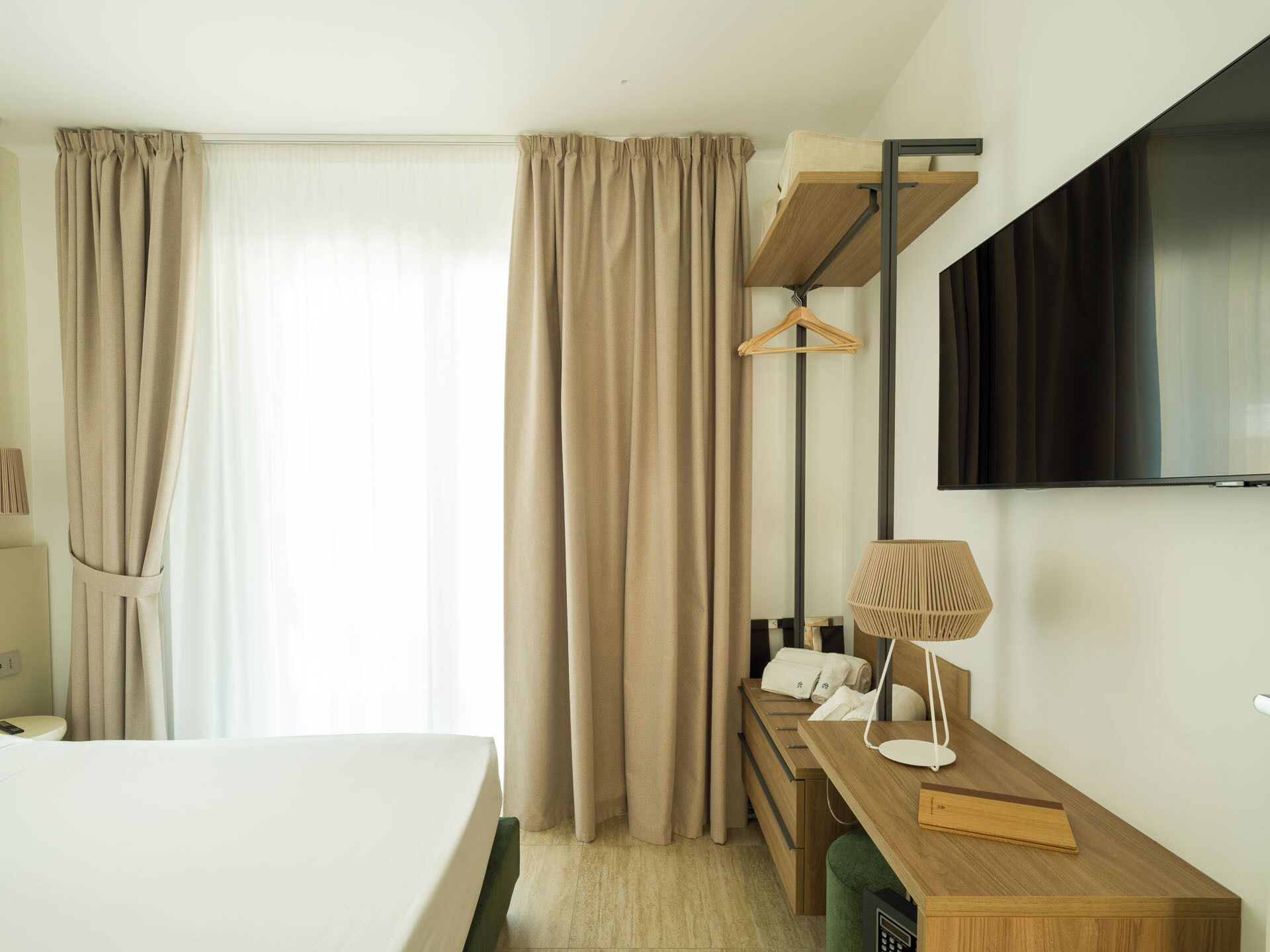 A hotel room with a bed, a desk with a lamp, a TV mounted on the wall, and curtains.