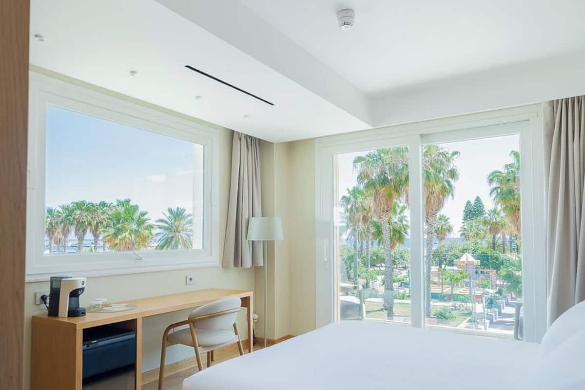 Quarto de hotel moderno com uma cama grande, uma escrivaninha com cadeira, e vista para palmeiras e um parque.