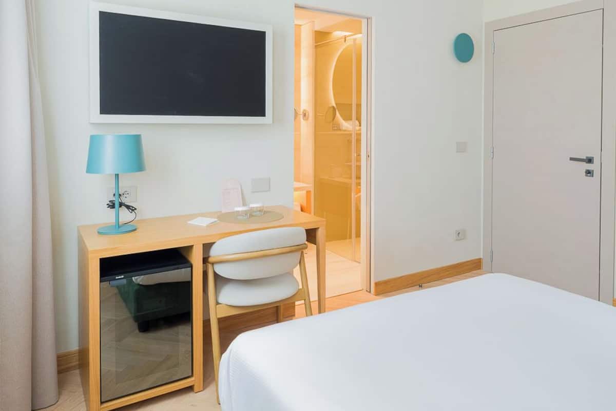 Quarto de hotel com cama, escrivaninha com luminária, televisão, guarda-roupa e banheiro com chuveiro.
