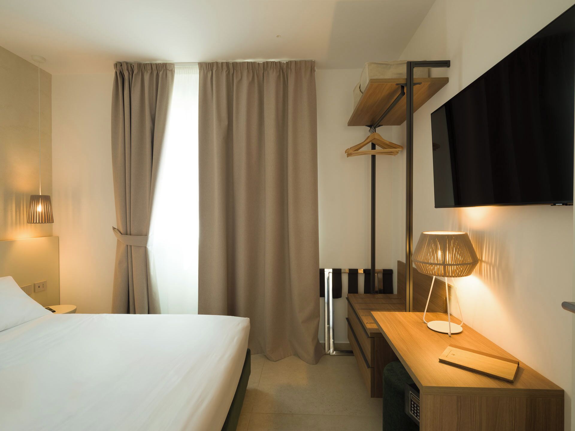 Una camera d'albergo con un letto, una scrivania, una sedia, un appendiabiti, una televisione e una lampada.