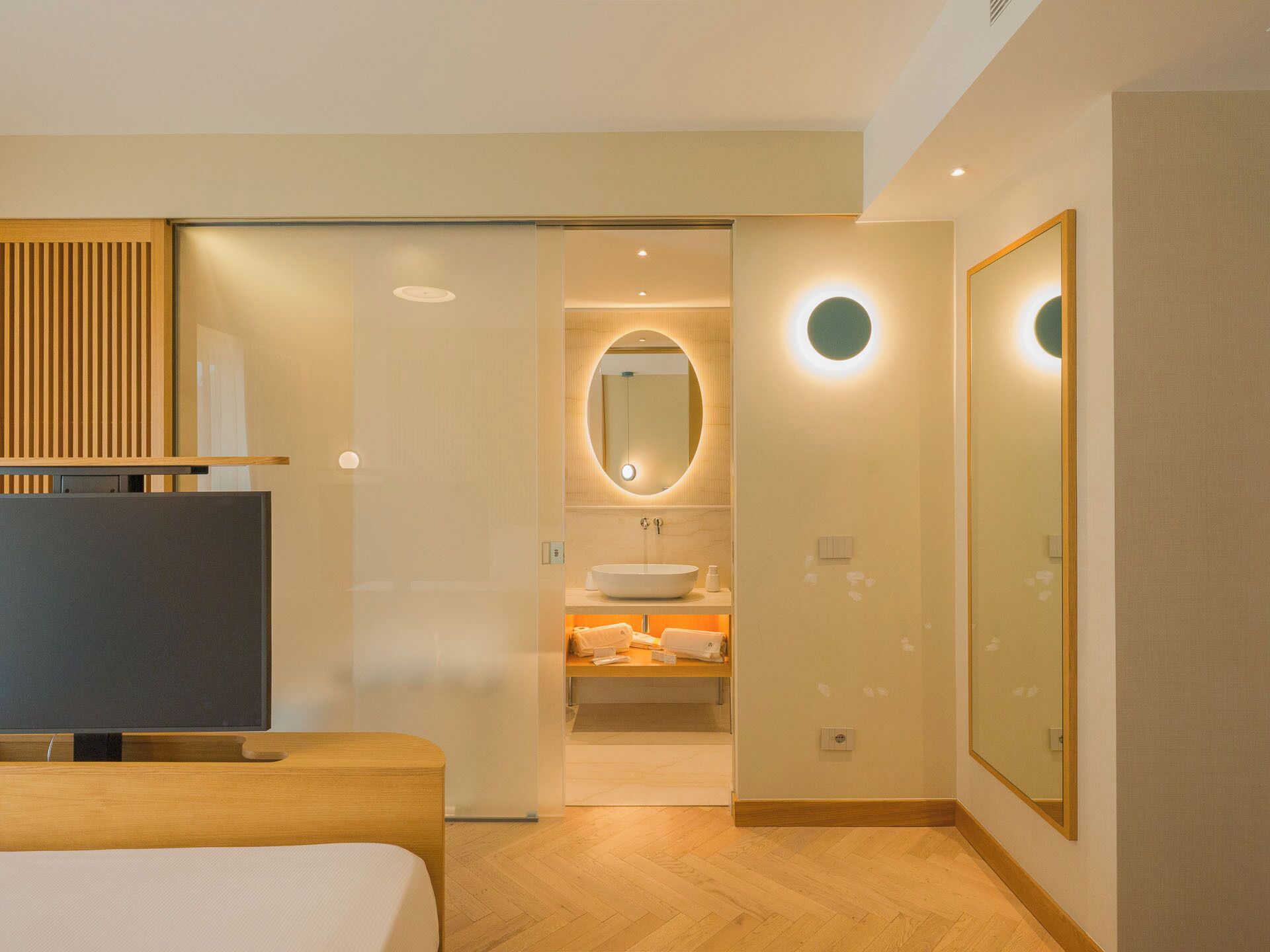 Een moderne hotelkamer met een bed, een televisie, een badkamer met een wastafel en spiegel, en een grote spiegel aan de muur.