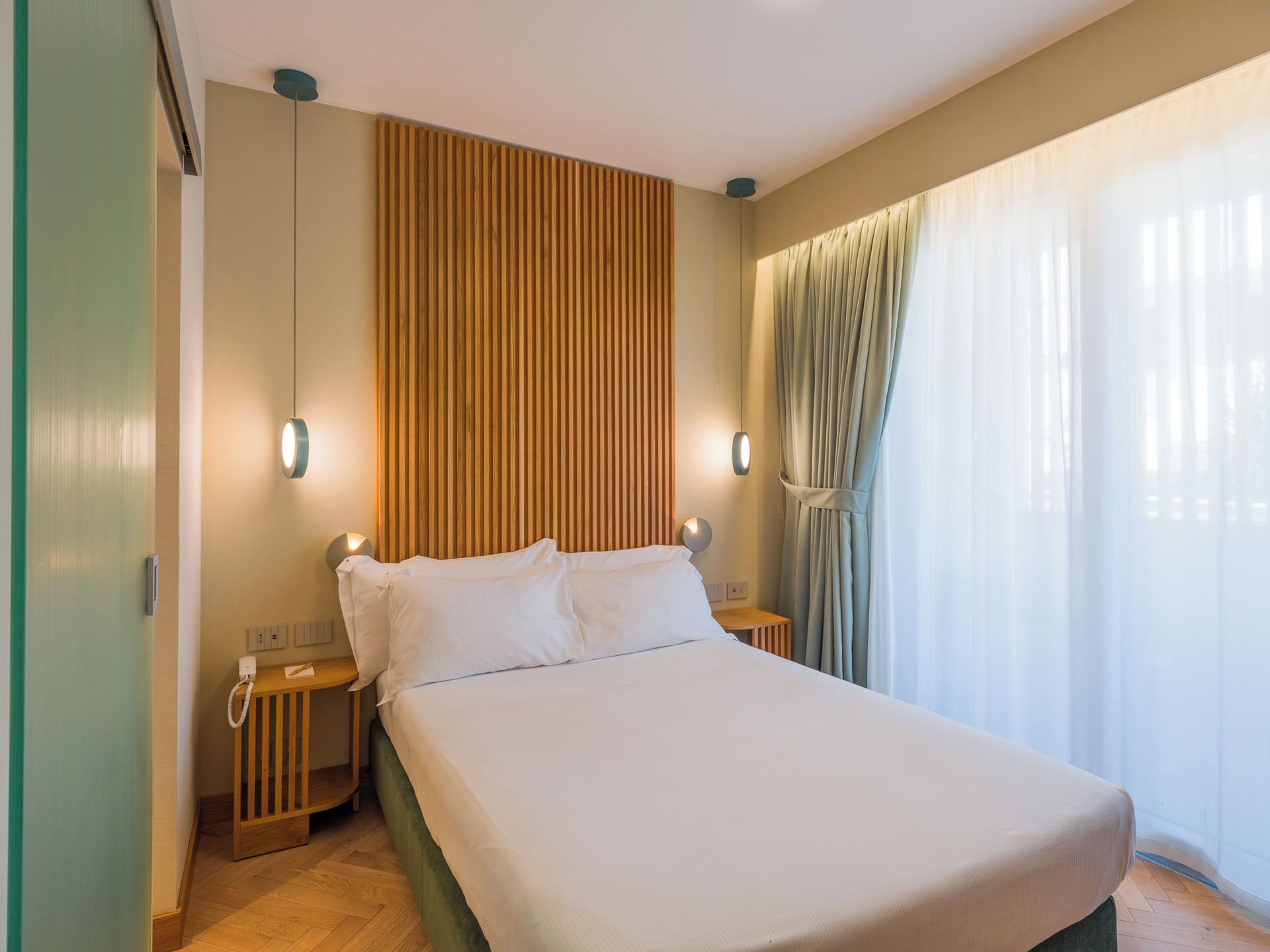 A hotel room with a large bed, wooden panel wall, two bedside tables with lamps, a telephone, and a window with curtains.