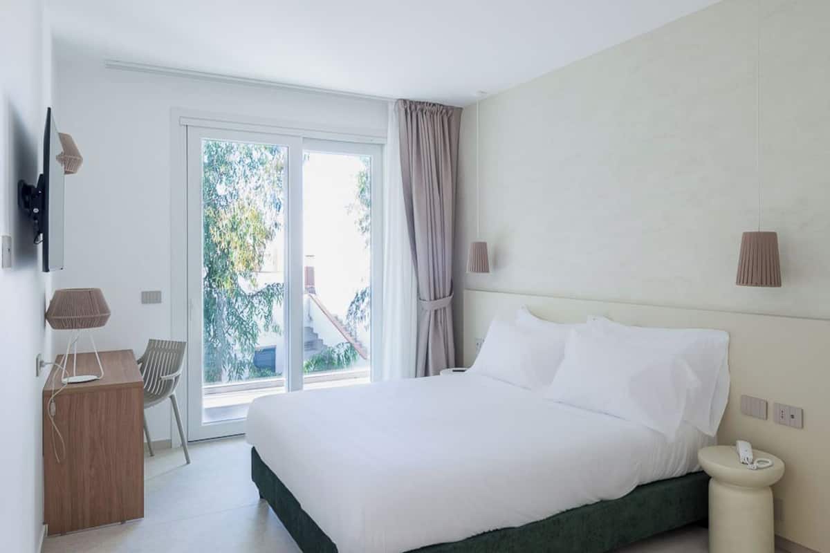Quarto moderno com cama grande, escrivaninha, cadeira e vista para o verde lá fora.