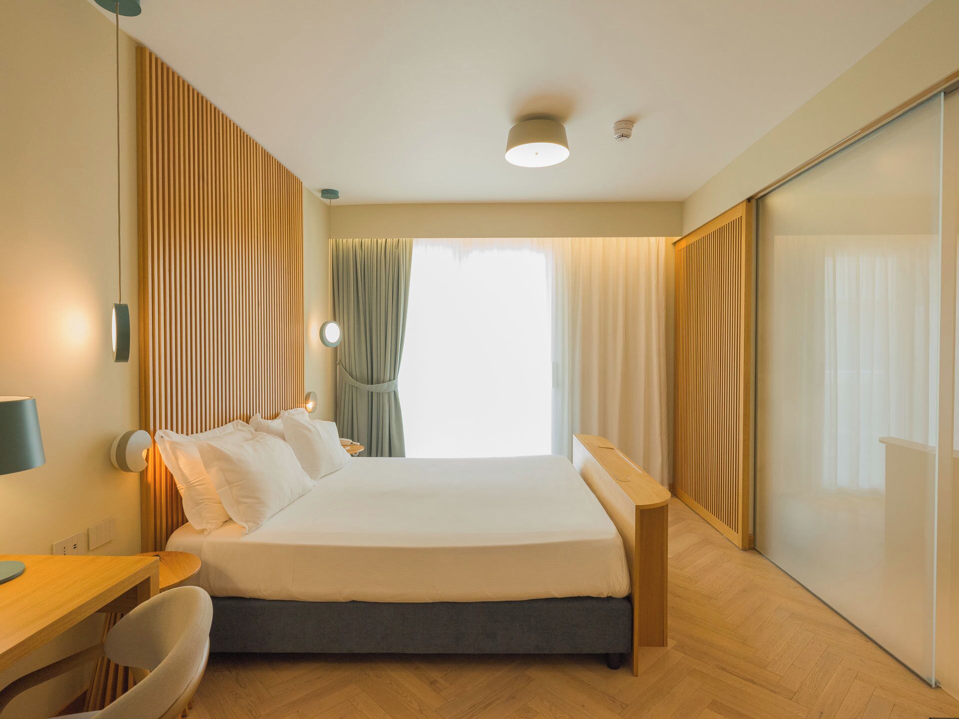 A modern hotel room with a bed, bedside table, lamp, and a view of the outside.