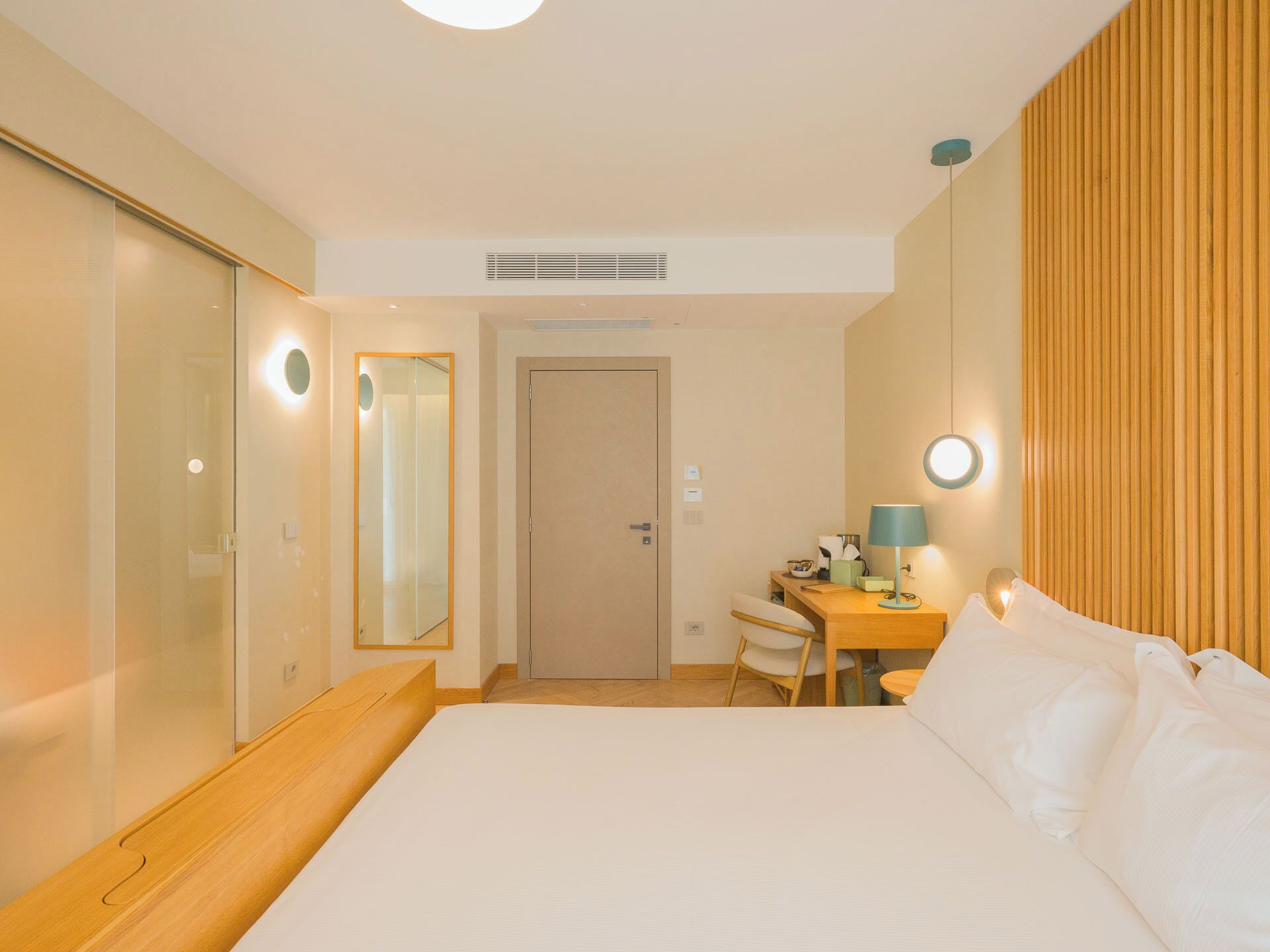 A hotel room with a bed, a desk, a chair, a mirror, and a door.
