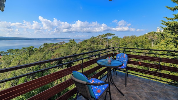 A balcony with a table and chairs, overlooking a scenic view of trees and a body of water. - Apartman Sea Blue  (Crikvenica)