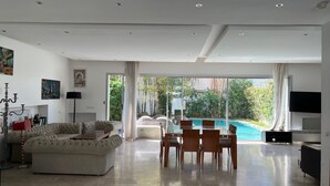 Un salon spacieux avec un grand canapé, une table à manger et des chaises, un téléviseur, et une vue sur une piscine et un espace verdoyant.