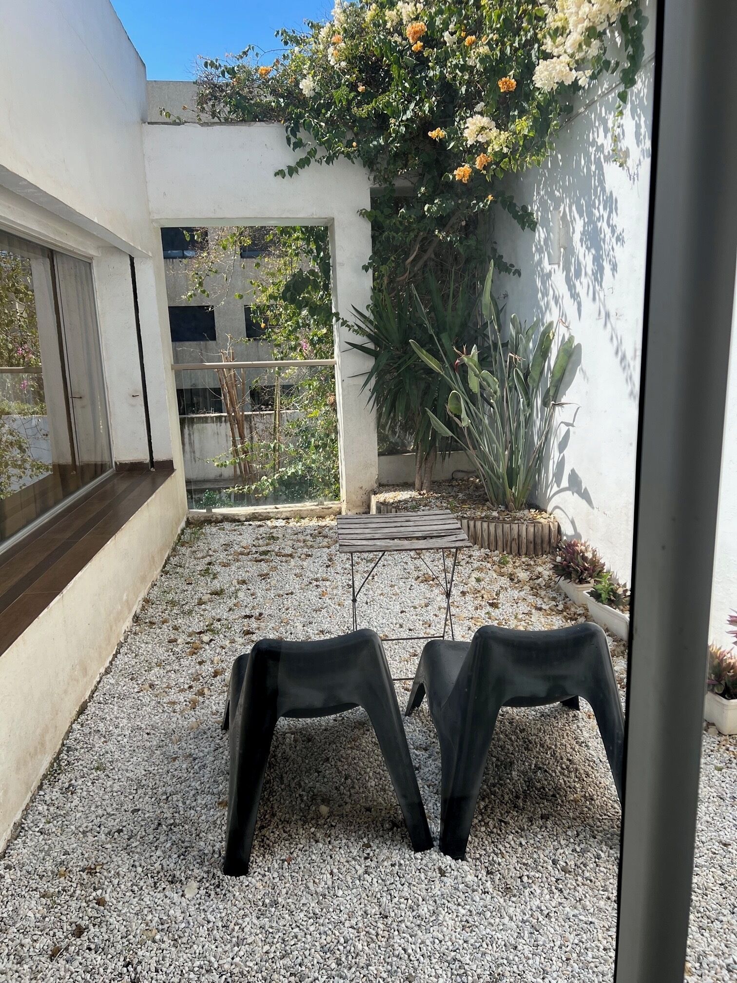 A patio area with a gravel floor, two black plastic chairs, a small table, and various potted plants.