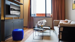 A modern living room with a flat-screen TV, a sofa, a coffee table, and a blue ottoman. - ipartment Koeln Oper @HOME by Best Western (Cologne)