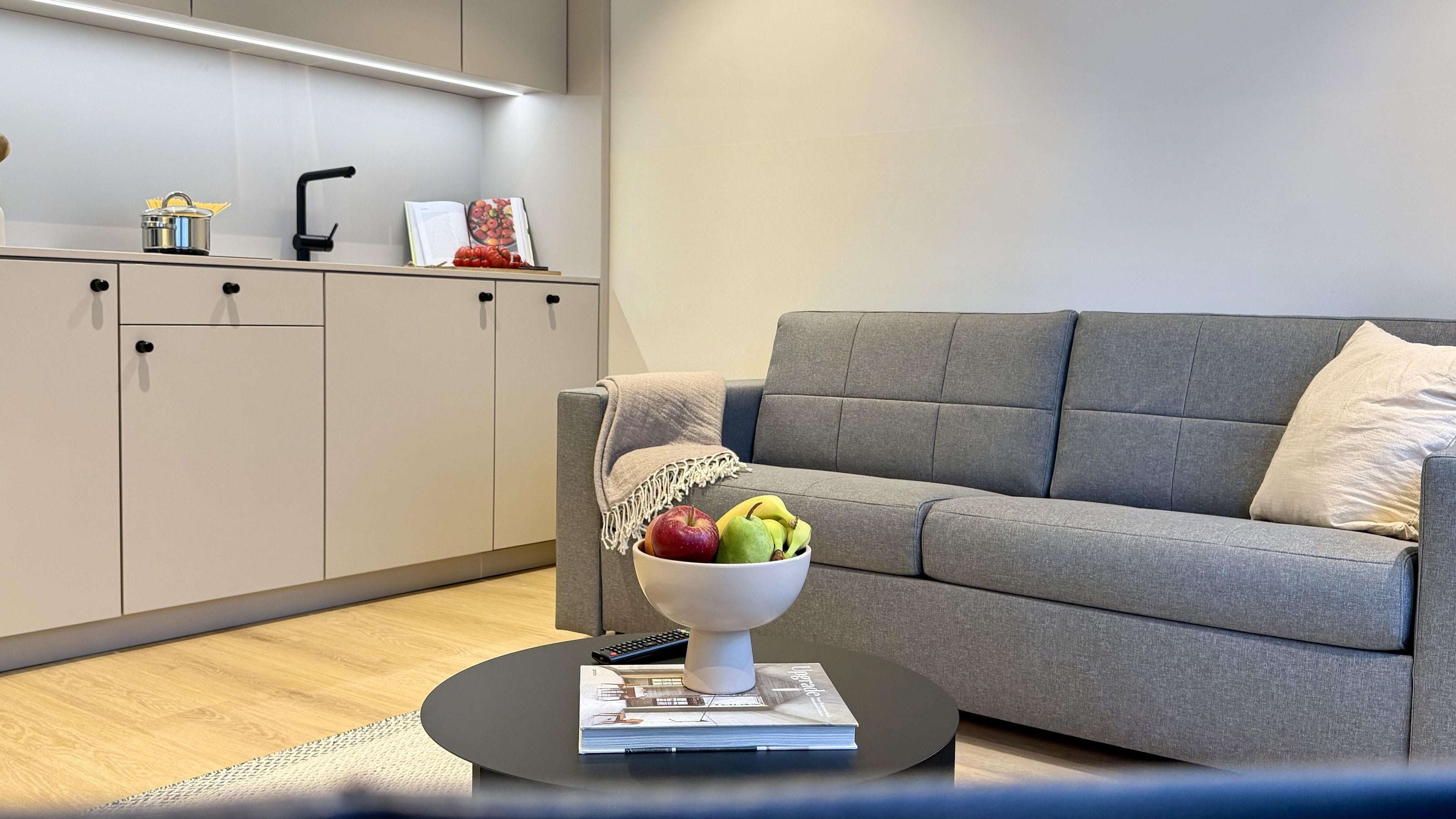 A modern living room with a grey sofa, a coffee table with a bowl of fruit, and a kitchen area with cabinets and a sink.