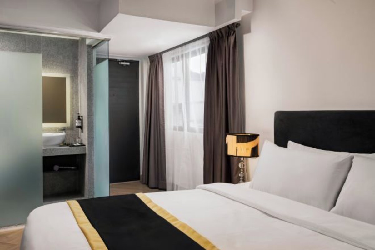 Una habitación de hotel con cama, mesita de noche con lámpara, baño con lavamanos y espejo, y vistas al exterior a través de una ventana con cortinas.