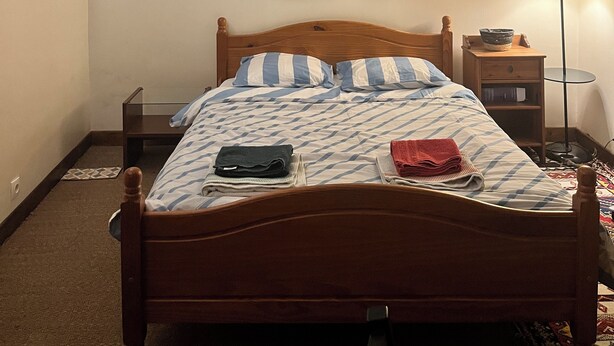 Quarto com cama de madeira, mesa de cabeceira, abajur e prateleira na parede.