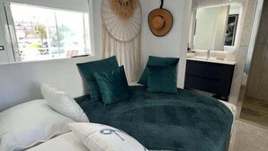 A modern bedroom with a large bed, two green pillows, a white pillow with a design, a wall-mounted hat, and a view of the outside through a window. - Lovely studio apartment next to the beach AV-63 (ROSES)