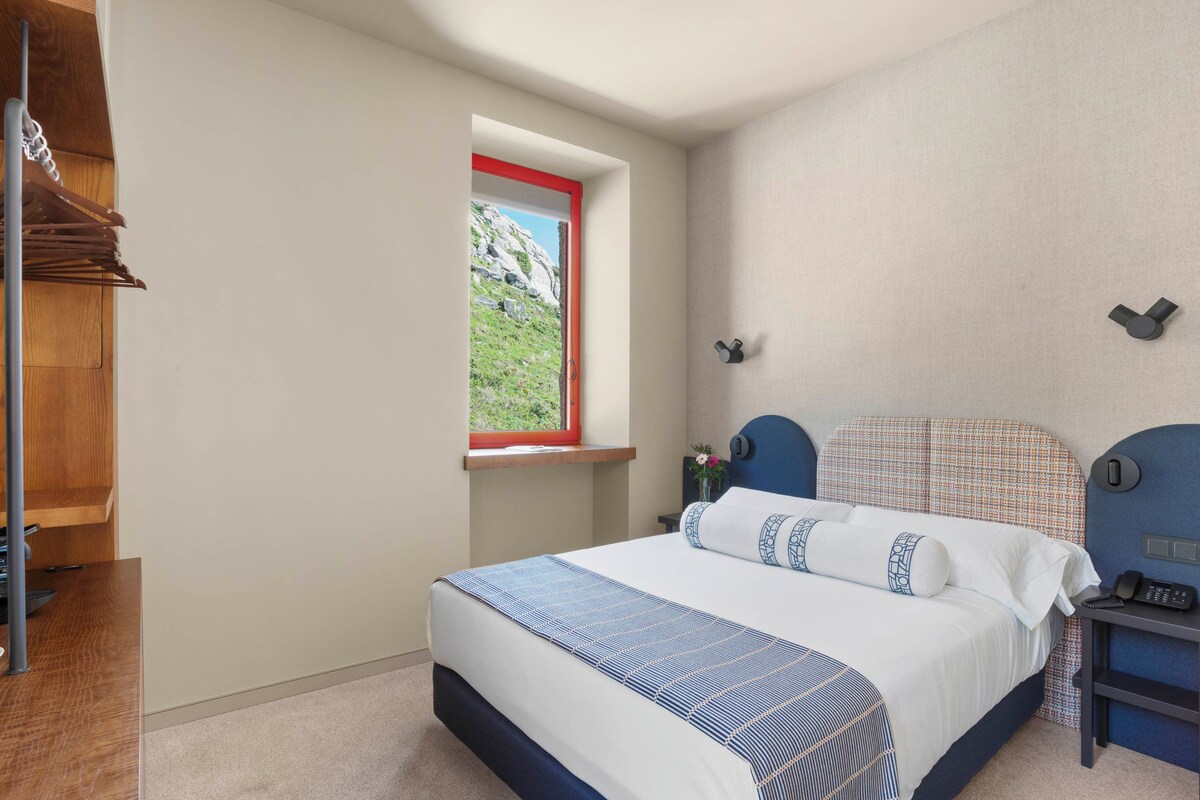 A bedroom with a bed, a nightstand, a window with a view of a rocky landscape, and a wooden shelf.