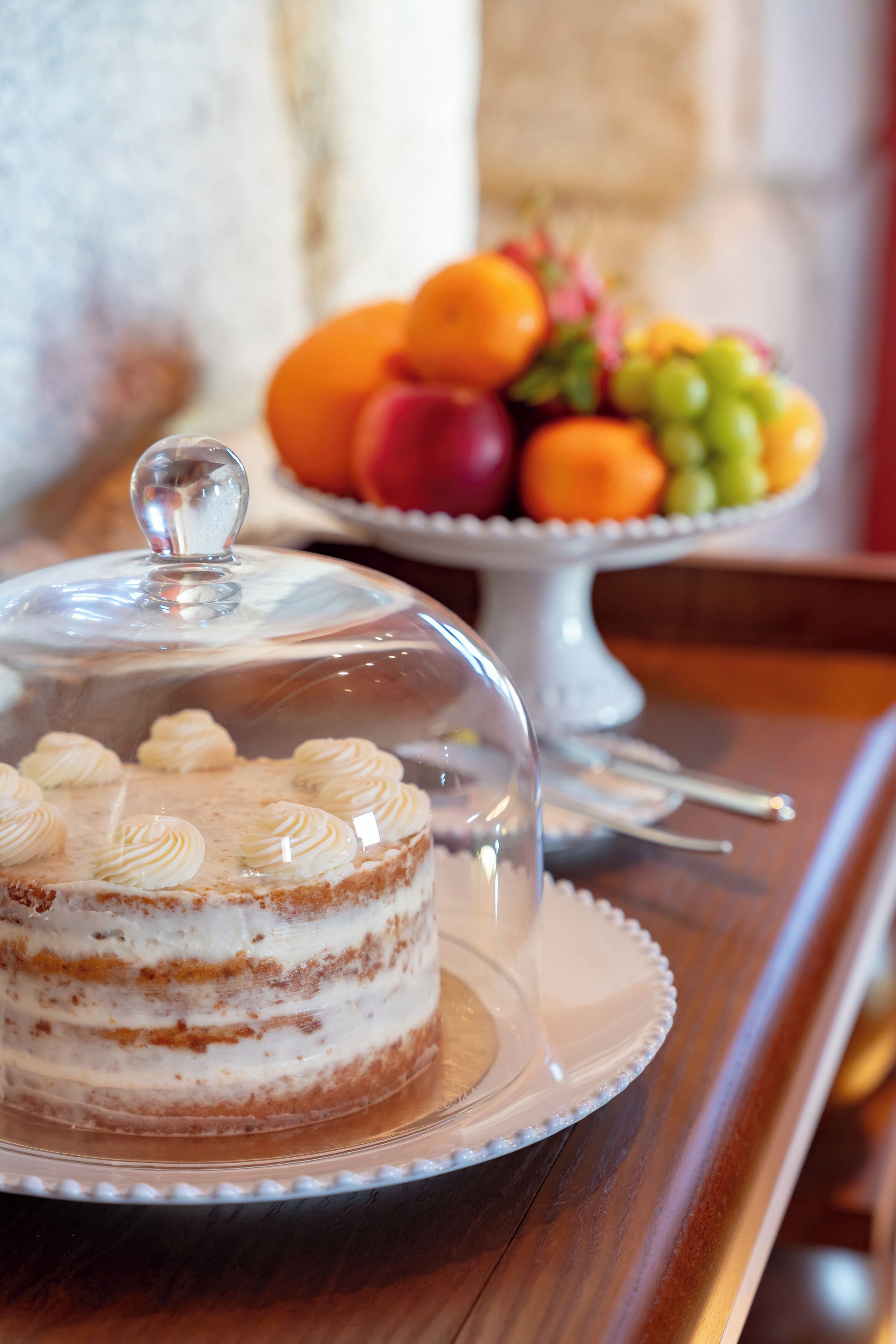 Un pastel con una campana de cristal sobre un plato blanco, una bandeja de frutas al fondo y una mesa de madera.
