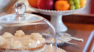 Un pastel con una campana de cristal sobre un plato blanco, una bandeja de frutas al fondo y una mesa de madera.