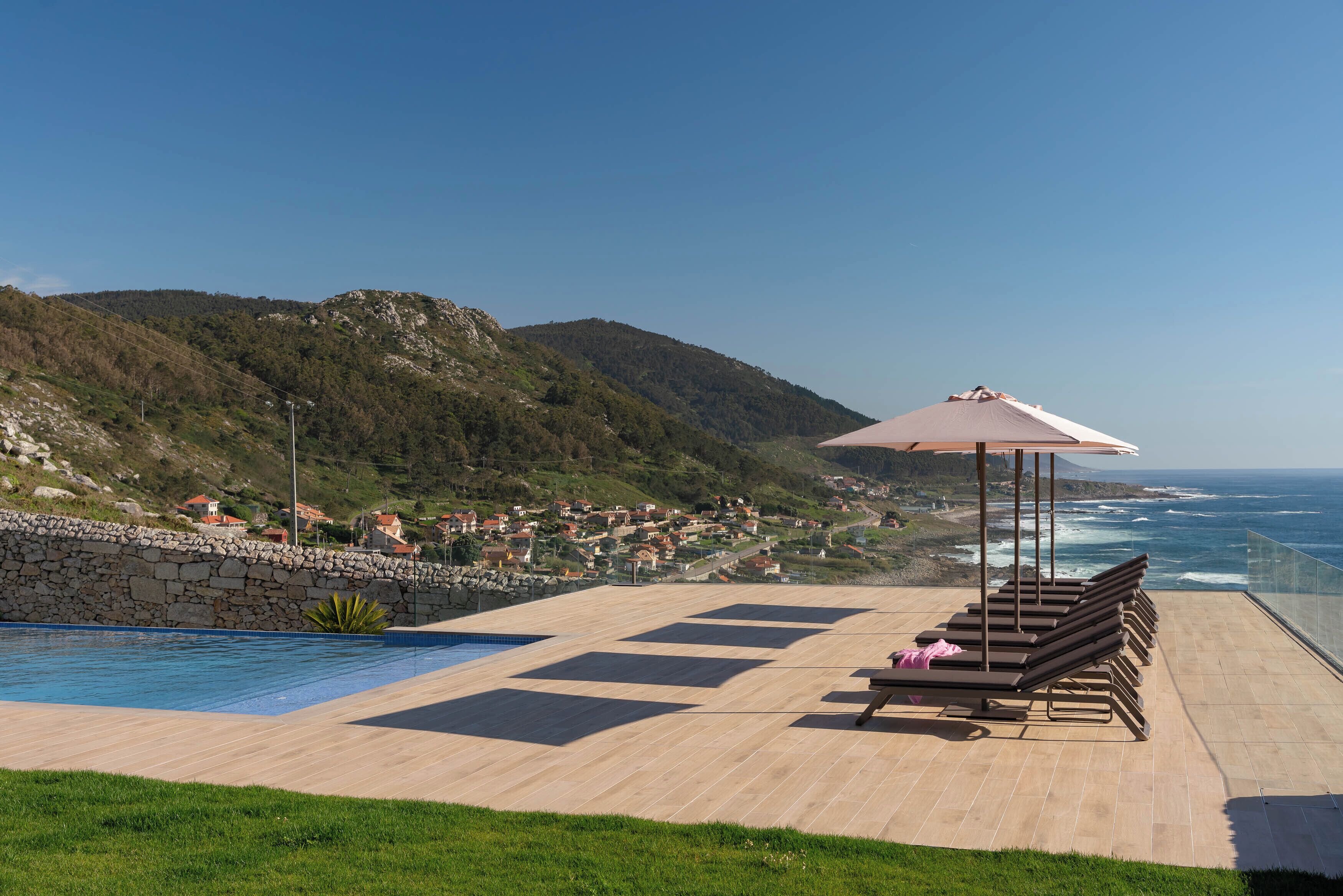 A poolside area with lounge chairs and an umbrella, overlooking a coastal town and ocean.