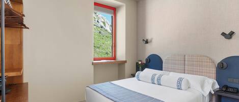 Un dormitorio con cama, mesita de noche, ventana con vista a un paisaje rocoso y estante de madera.