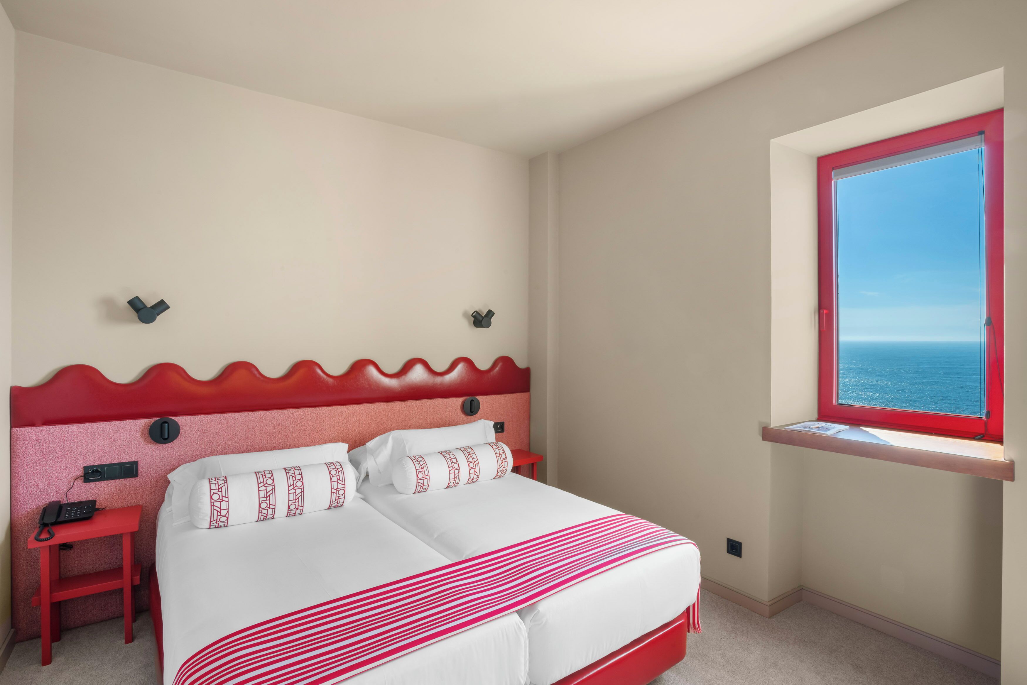 A hotel room with a bed, a small desk, a telephone, a window with a red frame, and a view of the sea.