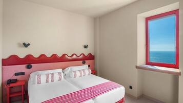 A hotel room with a bed, a small desk, a telephone, a window with a red frame, and a view of the sea.