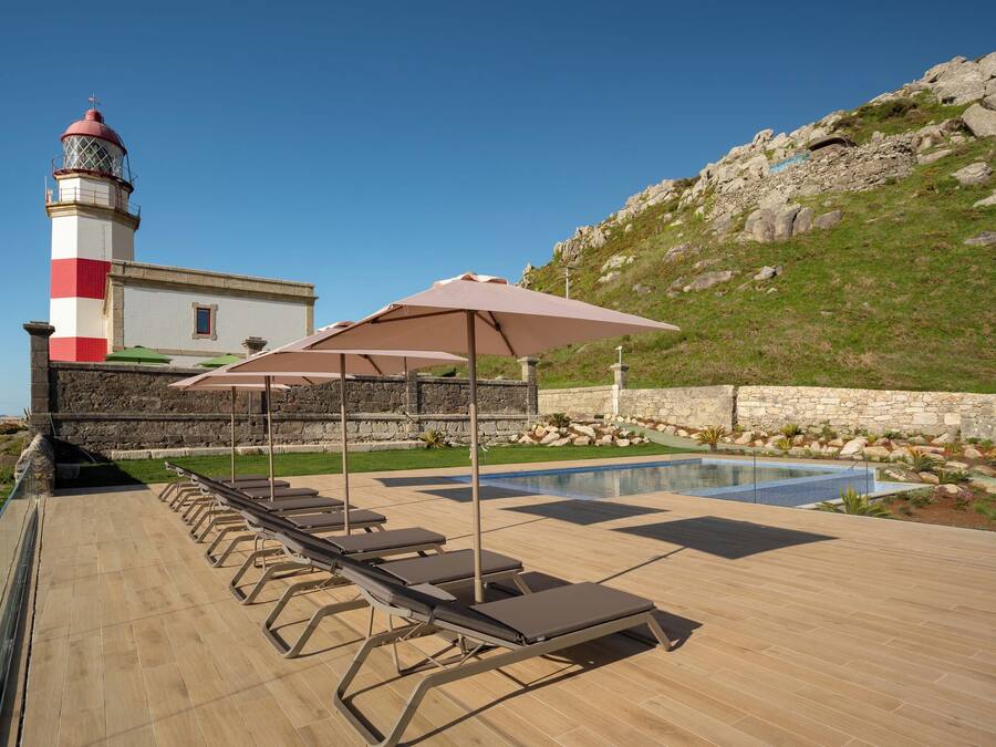 Un área de piscina con sillas de descanso y sombrillas, un muro de piedra y un faro al fondo.