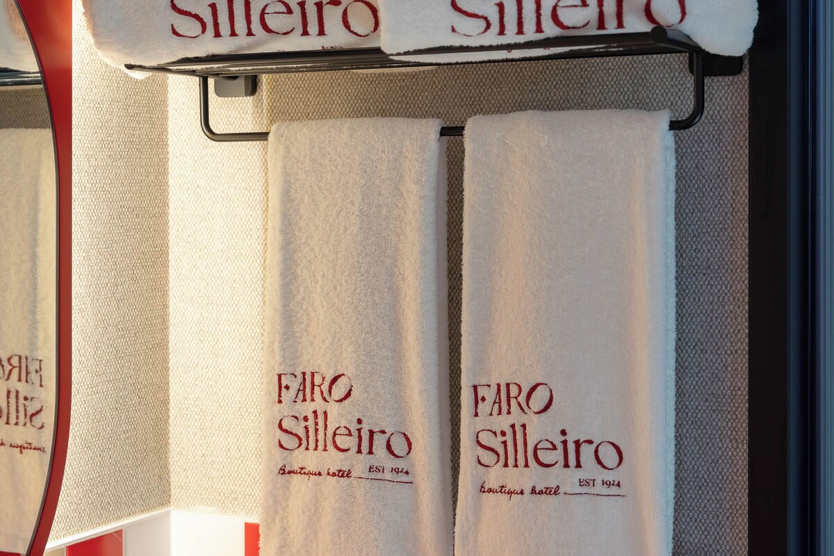 Un baño con un perchero que sostiene tres toallas blancas con la inscripción 'FARO Silleiro'.