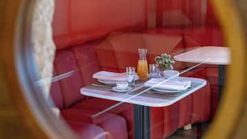 A circular window view of a red cushioned seating area with a table set for two, including a glass of orange juice and a small vase with flowers.
