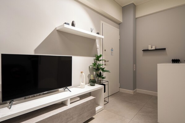 A modern living room with a flat-screen TV on a white console table, a potted plant, and a small shelf with decorative items.