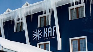 A building with a dark blue exterior, white trim, and icicles hanging from the roof. The building has multiple windows and a stone wall on the left side. - KAF MOUNT ERCİYES (Hacilar)