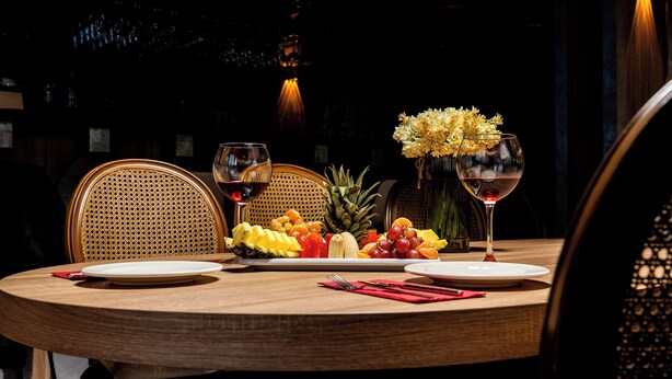 A round wooden table set for two with plates, cutlery, and a fruit arrangement. Two wine glasses and a vase with flowers are also visible. - KAF MOUNT ERCİYES (Hacilar)