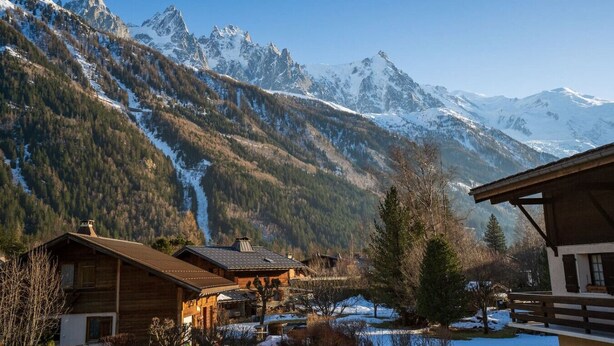 Eine Berghütte mit Holzhäuschen, einer Skipiste und schneebedeckten Gipfeln im Hintergrund.