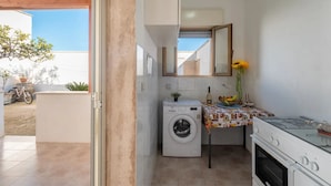 Une cuisine compacte équipée d’un lave-linge, d’une cuisinière et d’une petite table sur laquelle se trouvent un panier de fruits et un vase rempli de tournesols.