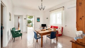 Un coin repas avec une table en bois, des chaises et un lustre. On y trouve un canapé avec un coussin rouge et une fenêtre avec des rideaux.