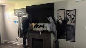 A living room with a fireplace, a flat-screen TV mounted above it, and decorative wall art. - Charming 3-bedroom house with WiFi and AC in West Monroe (West Monroe)