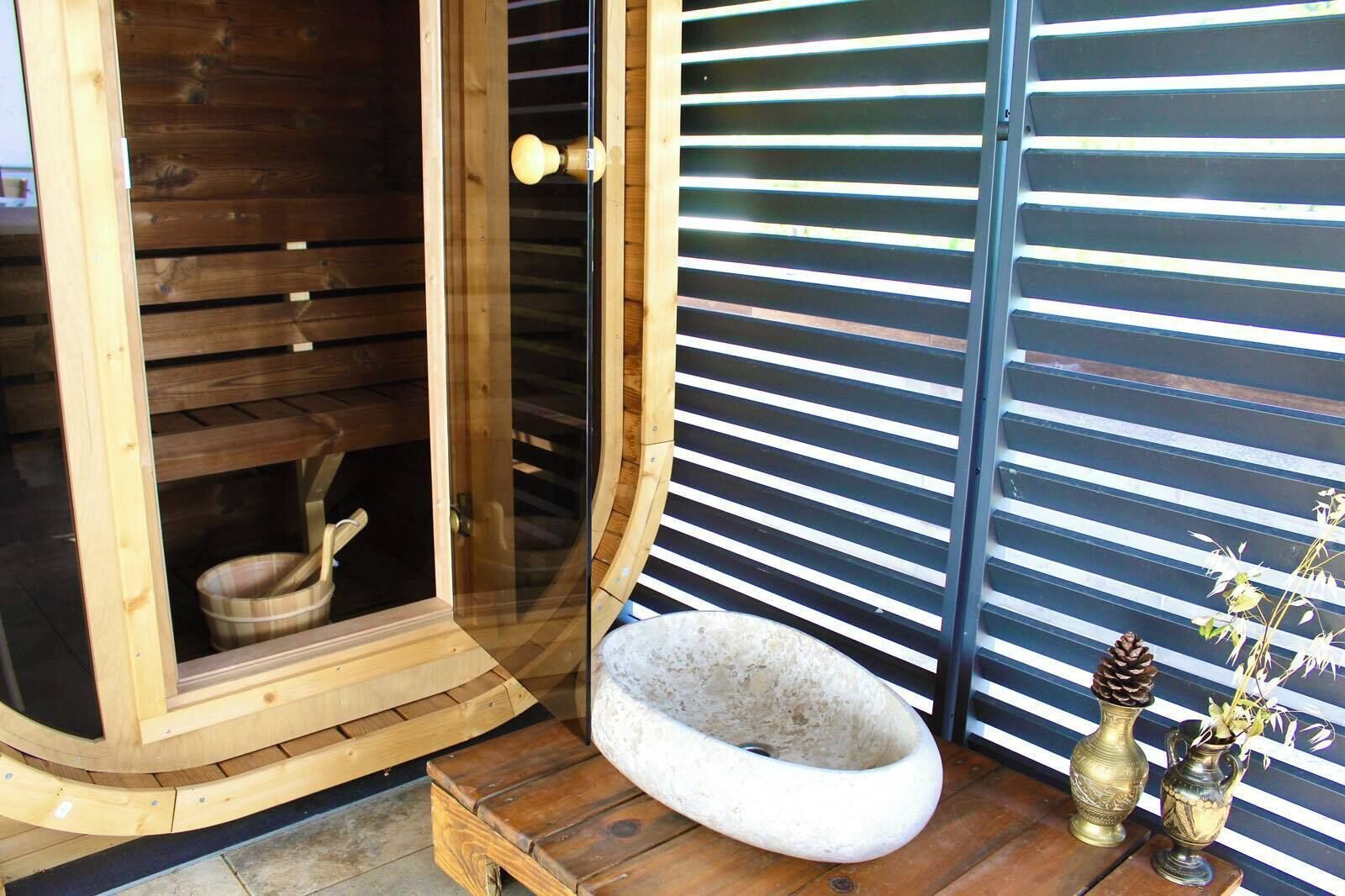 A wooden sauna with a glass door, a stone bench, and decorative vases on a wooden deck.