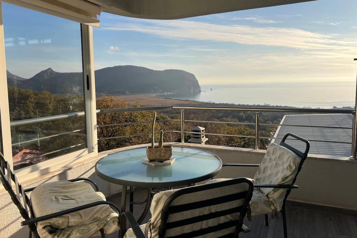 A balcony with a glass table and chairs, overlooking a coastal landscape with mountains and a body of water.
