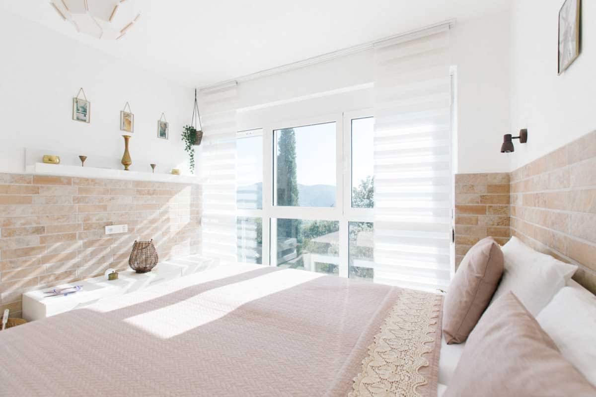 A bedroom with a large window, a brick fireplace, and a bed with pillows.