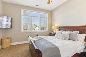 A bedroom with a large bed, a bedside table with a lamp, a TV mounted on the wall, and a window with blinds.