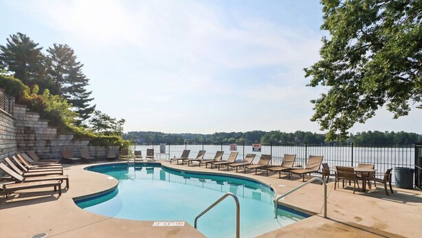 A swimming pool with lounge chairs and tables, surrounded by a stone wall and a fence, with a view of a body of water and trees in the background. - Penthouse Paradise at Delton Grand (Wisconsin Dells)