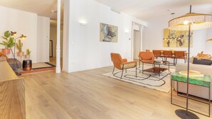 Una sala de estar moderna con piso de madera, una mesa de centro, sillas naranjas y un sofá. Hay un comedor con una mesa y sillas, y un cuadro grande en la pared.