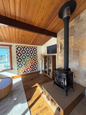 Une pièce chaleureuse avec un plafond en bois, un poêle à bois noir, une courtepointe aux couleurs vives et une vue par la fenêtre.