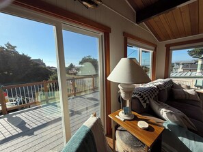 A room with a wooden table, a lamp, a sofa, and a view of a deck and outdoor area. - Welcome to Heron's Hearth (Cape Meares)