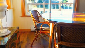 A room with a round glass table, two wicker chairs, a lamp, and a view of a wooden deck and trees outside.