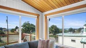 Una habitación con techo de madera, un sofá gris con cojines estampados, y ventanas amplias que dan a un edificio con tejado verde y árboles.