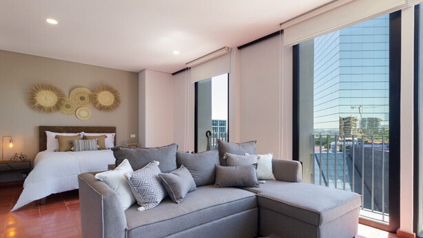Un salon moderne avec un canapé d’angle gris, une petite table basse et un lit recouvert d’une couette blanche.