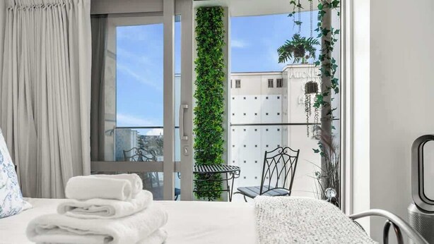 Una habitación de hotel con cama, una pila de toallas ordenadas, un ventanal con vistas a un edificio y follaje, y un balcón con una silla y una mesa.