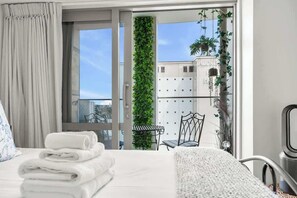 A hotel room with a bed, a neatly folded stack of towels, a window with a view of a building and greenery, and a balcony with a chair and a table.